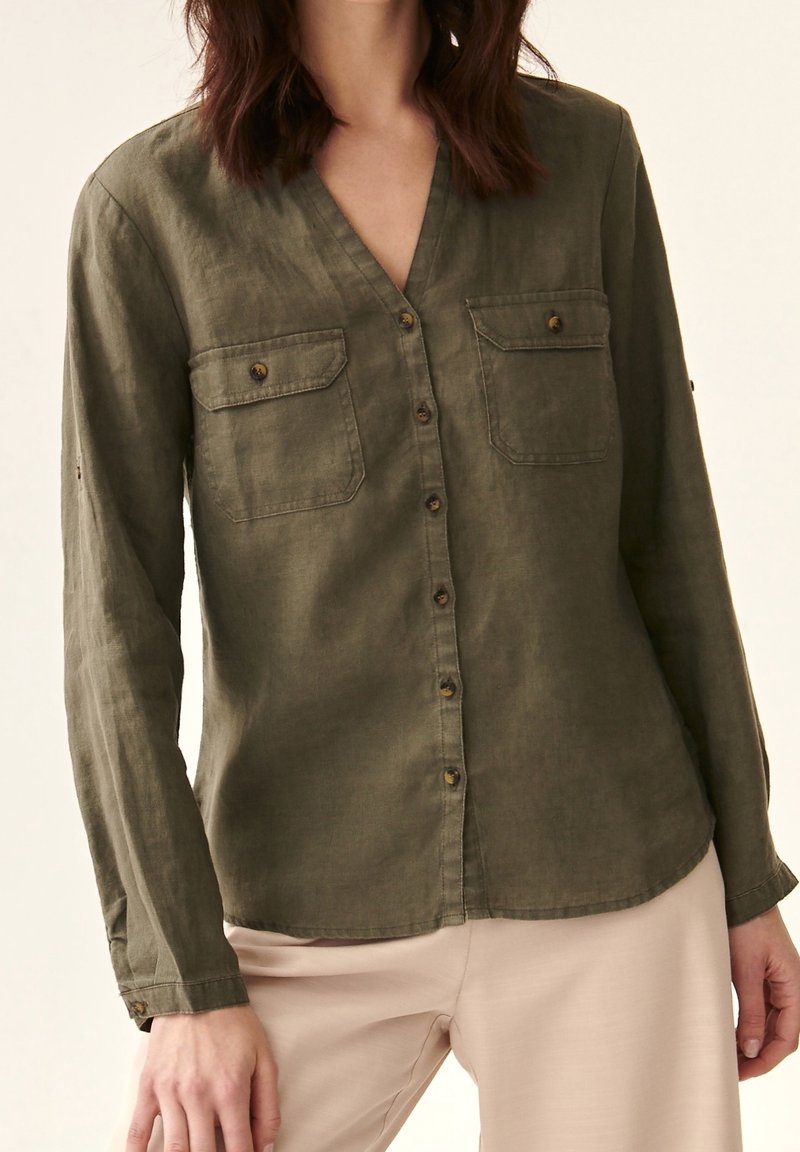 Femme portant une chemise vert olive à boutons avec deux poches poitrine et un pantalon beige, debout devant un fond uni.