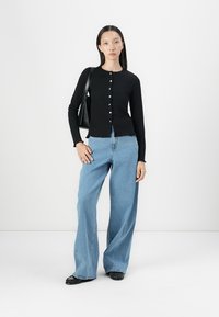 Zwarte geribbelde lange mouw top met knoopsluiting, gecombineerd met blauwe wijde jeans. Model houdt een zwarte schoudertas vast.