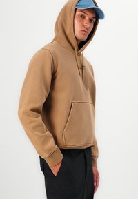 Brown hooded sweatshirt with a front pocket, cotton fabric, ribbed cuffs, and adjustable drawstring. Worn with black pants and a light blue cap.