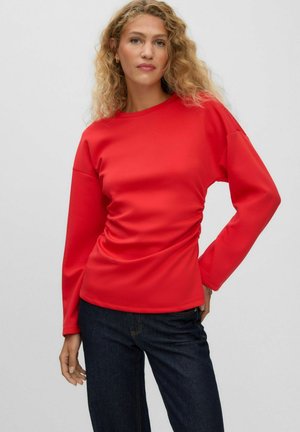 Femme aux cheveux blonds bouclés portant un haut à manches longues rouge vif et un jean bleu foncé, debout devant un fond clair uni.