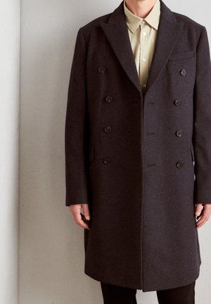 Mand iført en mørkebrun dobbeltradet frakke over en lysebrun skjorte med knapper, stående mod en ensfarvet væg.