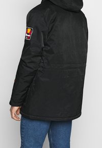 Chaqueta negra aislante con capucha, que presenta un parche en la manga izquierda y una textura suave y brillante. El diseño es minimalista.