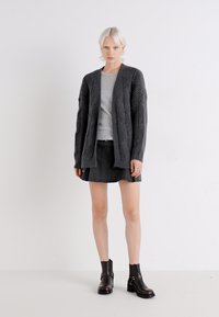 Dunkelgrauer Strickcardigan mit gerippter Textur und offenem Vorderteil, kombiniert mit einem hellgrauen Top und einem schwarzen Rock. Schwarze Ankle-Boots runden den Look ab.