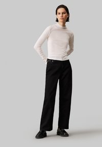 Haut long à manches blanches avec col montant, associé à un jean noir taille haute à larges jambes et des chaussures à lacets noires. Textures lisses et design ajusté.