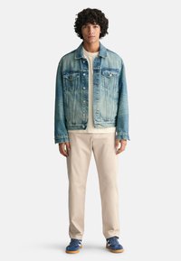 Hellblaue Jeansjacke mit zwei Brusttaschen, Knopfverschluss und ausgewaschenem Look. Kombiniert mit beigen Hosen und blauen Turnschuhen.