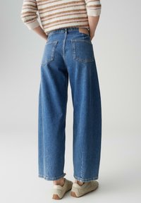 Jean bleu à jambes larges avec une taille haute, deux poches arrière, et une étiquette de marque en cuir à l'arrière. Associé à des baskets gris clair.