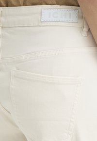 Gros plan sur la poche arrière d'un jean en denim blanc avec une étiquette de marque cousue "ICHI" sur la ceinture, sur un fond beige.
