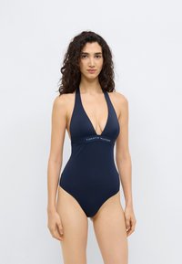 Μπλε ναυτικό ολόσωμο μαγιό με λαιμόκοψη halter και βαθύ V, με αντίθετη λευκή και κόκκινη ταινία με το λογότυπο "TOMMY HILFIGER" στη μέση.