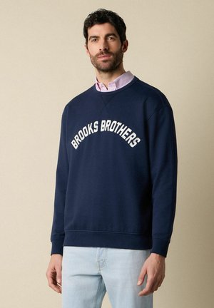Man draagt een marineblauwe sweatshirt met de tekst "Brooks Brothers" en lichtblauwe jeans, staand tegen een beige achtergrond.