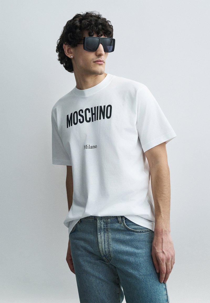 Biała bawełniana koszulka z nadrukiem „MOSCHINO” i „Milano” w czerni. Połączona z jasnoniebieskimi dżinsami. Modelka nosi duże czarne okulary przeciwsłoneczne.