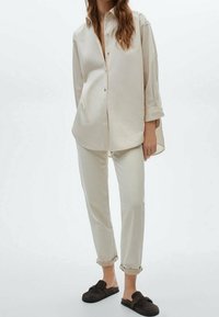 Chemise longue à manches longues beige avec boutons, coupe ample, associée à un pantalon beige clair retroussé aux chevilles et des chaussures à enfiler noires.