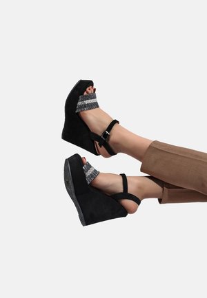 Pieds portant des sandales compensées noires avec des brides pailletées sur les orteils et des sangles à la cheville, assorties à un pantalon marron sur un fond uni.