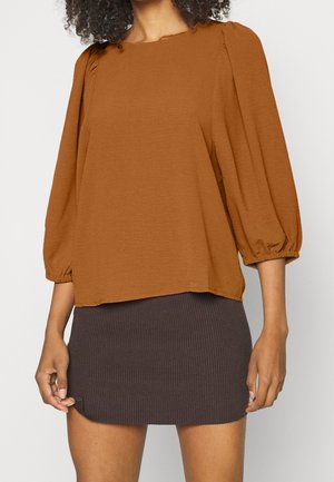 Blusa di colore ruggine, comoda e leggermente svasata, con maniche a sbuffo e dettagli con bottoni sul colletto, abbinata a una mini gonna a coste scura.