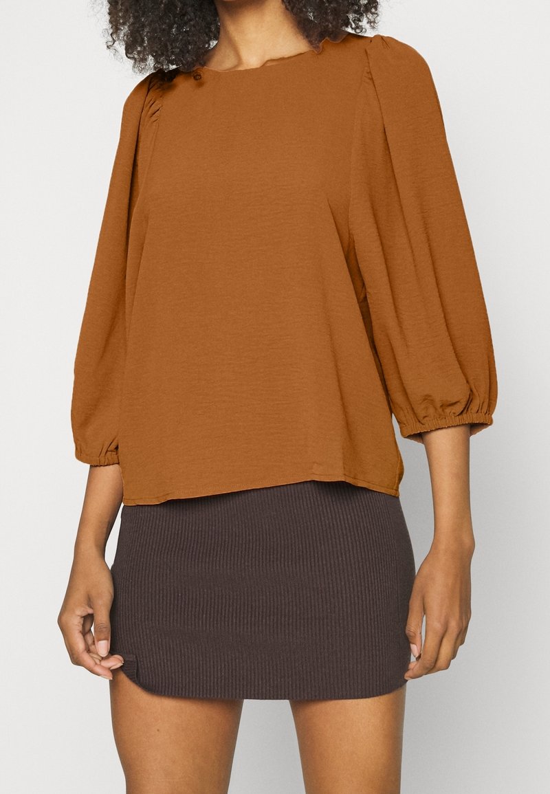 Blusa di colore ruggine, comoda e leggermente svasata, con maniche a sbuffo e dettagli con bottoni sul colletto, abbinata a una mini gonna a coste scura.