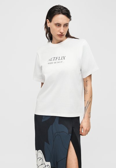 Γυναίκα με τατουάζ στο χέρι, που φοράει λευκό πουκάμισο με το κείμενο «Το Netflix με έκανε να το κάνω» και μαύρη φούστα με φλοράλ σχέδιο και εμπρόσθια σχισμή.