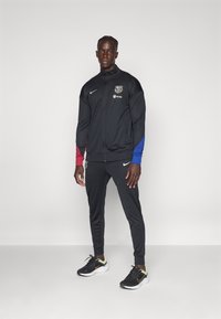 Sort atletiktøjssæt med farvede ærme accenter i rød og blå. Indeholder Nike-logo og Spotify-branding. Bæres med sorte sneakers.
