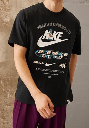 Personne portant un t-shirt Nike noir avec un texte sur la nature, l'équilibre et un slogan coloré en lettres bleues et oranges.
