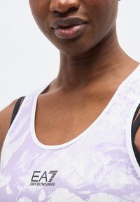 Κοντινό πλάνο ενός ατόμου που φορά αμάνικο τοπ σε ανοιχτό μοβ και λευκό με το λογότυπο "EA7 Emporio Armani" στο στήθος.