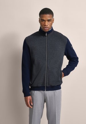 Grau strukturierte Zip-Jacke mit marineblauen Ärmeln, hohem Kragen und geripptem Saum; kombiniert mit grauen Hosen.