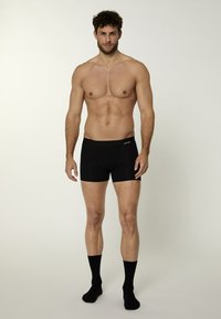 Boxer briefs neri con cintura elastica, su uno sfondo neutro. Modello che indossa calzini neri. Tessuto liscio, design aderente.
