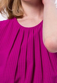 Donna che indossa una blusa magenta con pieghe vicino al colletto, sollevando il braccio sinistro vicino al volto, con capelli biondi e pelle chiara.