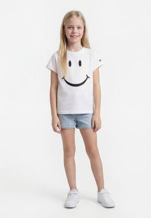 Jeune fille aux longs cheveux blonds portant un t-shirt blanc avec un grand smiley noir, un short en jean et des baskets blanches, souriante.
