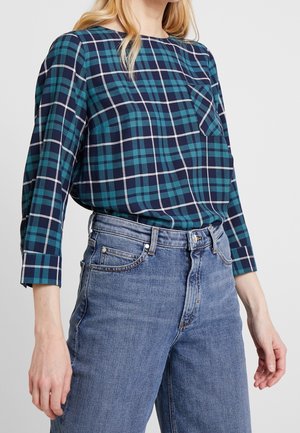 Geruit shirt in teal en marineblauw met witte accenten, ronde halslijn, lange mouwen en een borstzak, gecombineerd met lichtblauwe jeans.