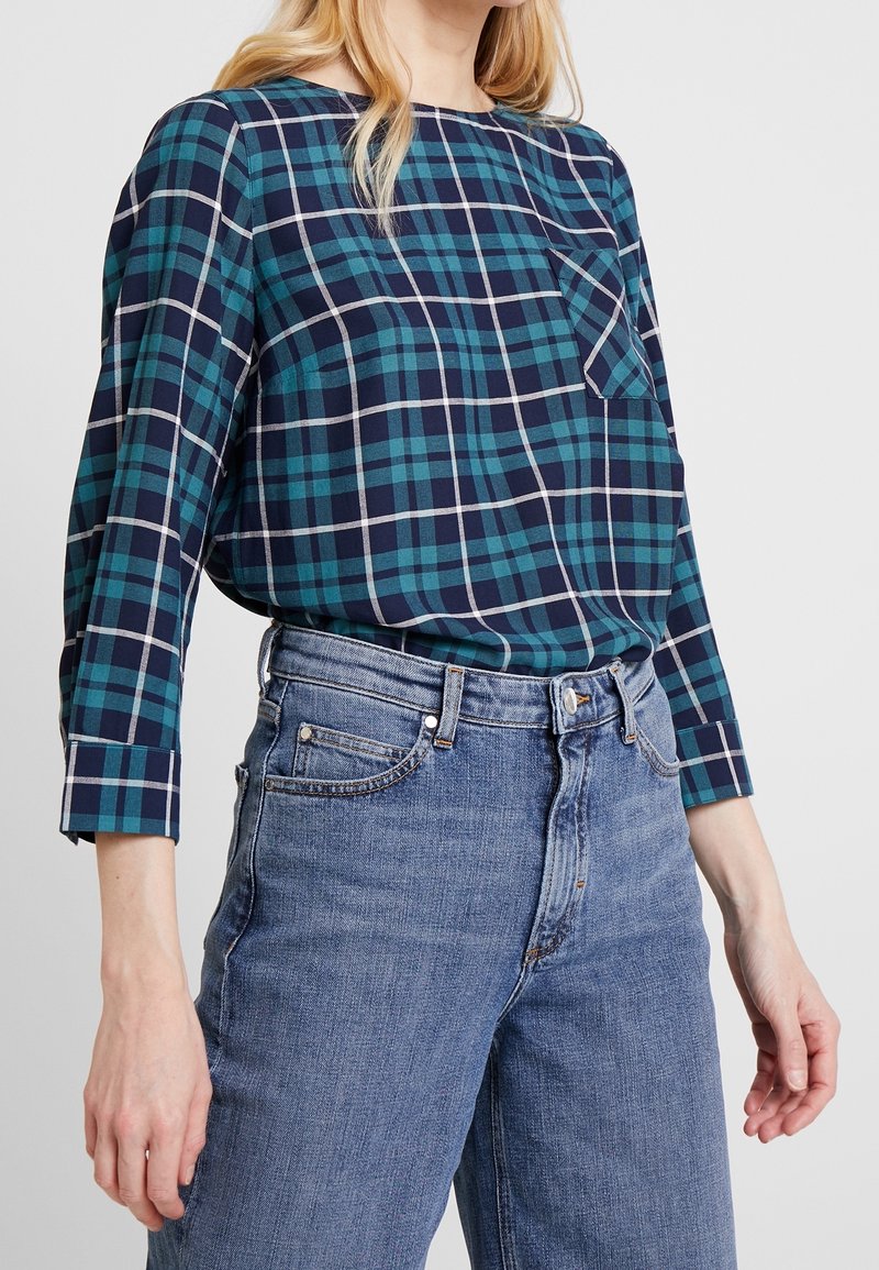 Chemise à carreaux en teal et bleu marine avec des accents blancs, col rond, manches longues et une poche poitrine, associée à un jean bleu clair.