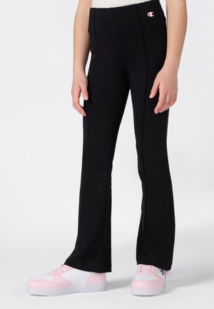 Enfant portant un pantalon noir évasé avec des coutures visibles et le logo Champion, associé à des baskets blanches et roses, se tenant sur un fond blanc.