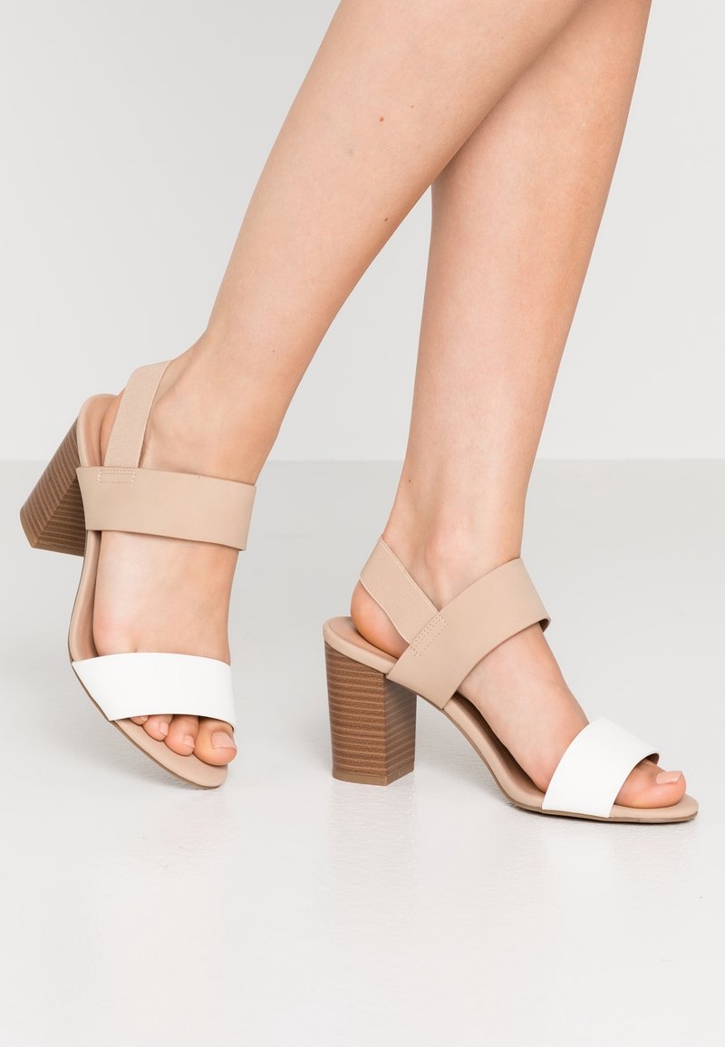 Pieds portant des sandales à talons blocs beige et blanches à bout ouvert avec brides à la cheville, sur un sol clair et un fond uni.