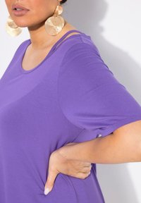 Lila kortärmad skjorta gjord av mjukt tyg, med rundad halsringning och en avslappnad passform. Stora strukturerade guldörhängen kompletterar outfiten.