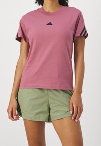 Růžové bavlněné tričko s černým logem hory, krátké rukávy s černými pruhy; v kombinaci s olivově zelenými šortkami, volný střih.