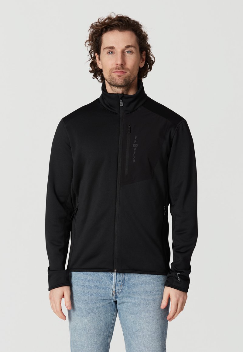 Schwarze Softshell-Jacke mit hohem Kragen, Reißverschluss vorne, Brusttasche und Seitentaschen; hat eine glatte, strukturierte Oberfläche.