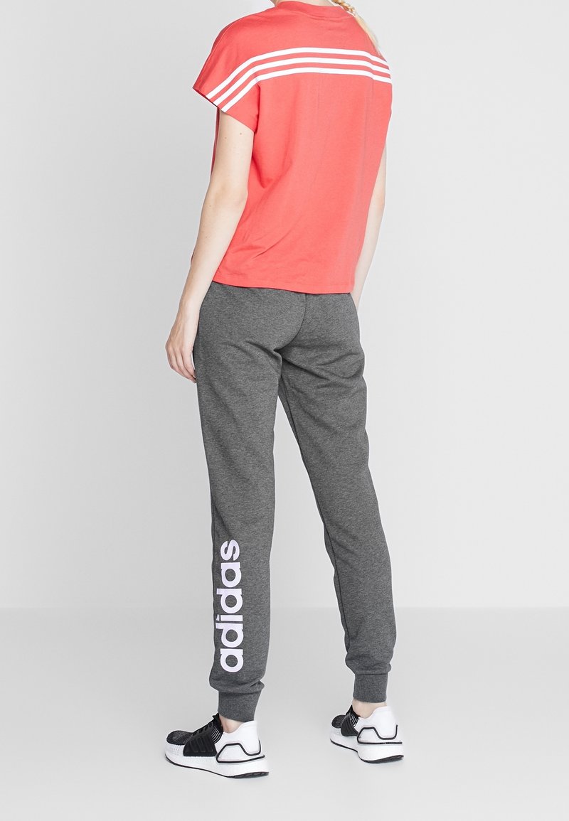 Koraalroze T-shirt met witte gestreepte accenten, gecombineerd met grijze joggingbroek met witte "adidas"-branding langs het been en zwarte sneakers.