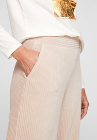 Pantalon en velours côtelé rose pâle à jambes larges, avec une texture côtelée verticale et des poches latérales. La ceinture est légèrement plissée.