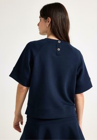 Námořnická modrá mikina s krátkým rukávem, kulatým výstřihem a dvěma zlatými knoflíky na zadní straně. Měkká textura a pohodlný střih.