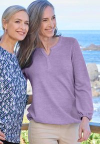 Zwei Frauen stehen draußen zusammen. Eine trägt ein lila Langarmoberteil mit einem Henley-Ausschnitt; die andere hat eine gemusterte Bluse mit lila Akzenten an.