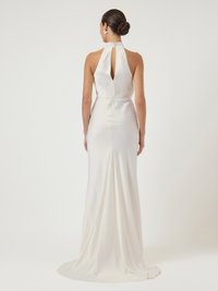 Γυναίκα με μαλλιά πιασμένα σε κότσο, φορώντας ένα αμάνικο άσπρο σατέν φόρεμα με κλειστό πίσω μέρος και μακριά ουρανό, στέκεται μπροστά σε ανοιχτό φόντο.