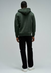 Sudadera con capucha de color verde oscuro y corte relajado, combinada con pantalones negros. El atuendo se complementa con zapatillas blancas.