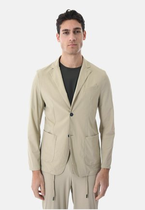 Giovane uomo che indossa una giacca beige su misura sopra una camicia nera con pantaloni beige abbinati, in piedi contro uno sfondo chiaro e uniforme.