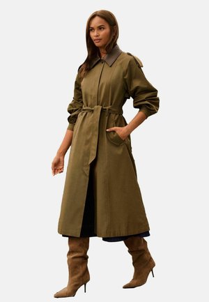 Kvinde iført en olivengrøn trenchcoat med bælte, mørkraver og brune knæhøje støvler med hæl, stående med den ene hånd i lommen.