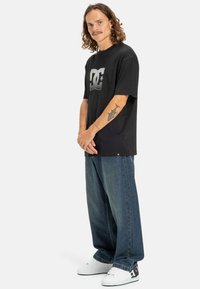 Black short-sleeve t-shirt featuring a large gray DC logo, paired with loose blue denim jeans and white sneakers.