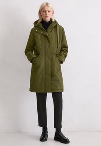 Olivgrön jacka knälång med huva, framknappar, två sidofickor och ribbade muddar. Bärs över svarta byxor och skor.