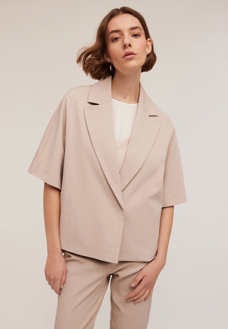 Blazer de gran tamaño en beige claro con solapa, mangas cortas y cierre frontal, combinado con una blusa blanca y pantalones a juego.