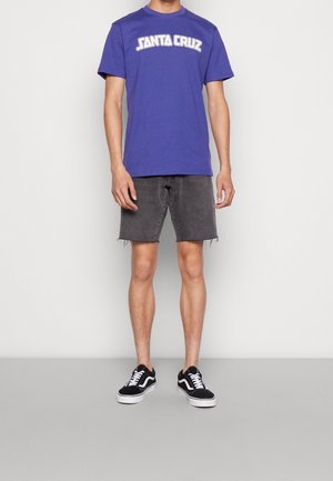 Violetti lyhythihainen t-paita, jossa on "SANTA CRUZ"-kuvio, yhdistetty harmaisiin farkkushortseihin ja mustiin Vans-tennareihin valkoisilla yksityiskohdilla.