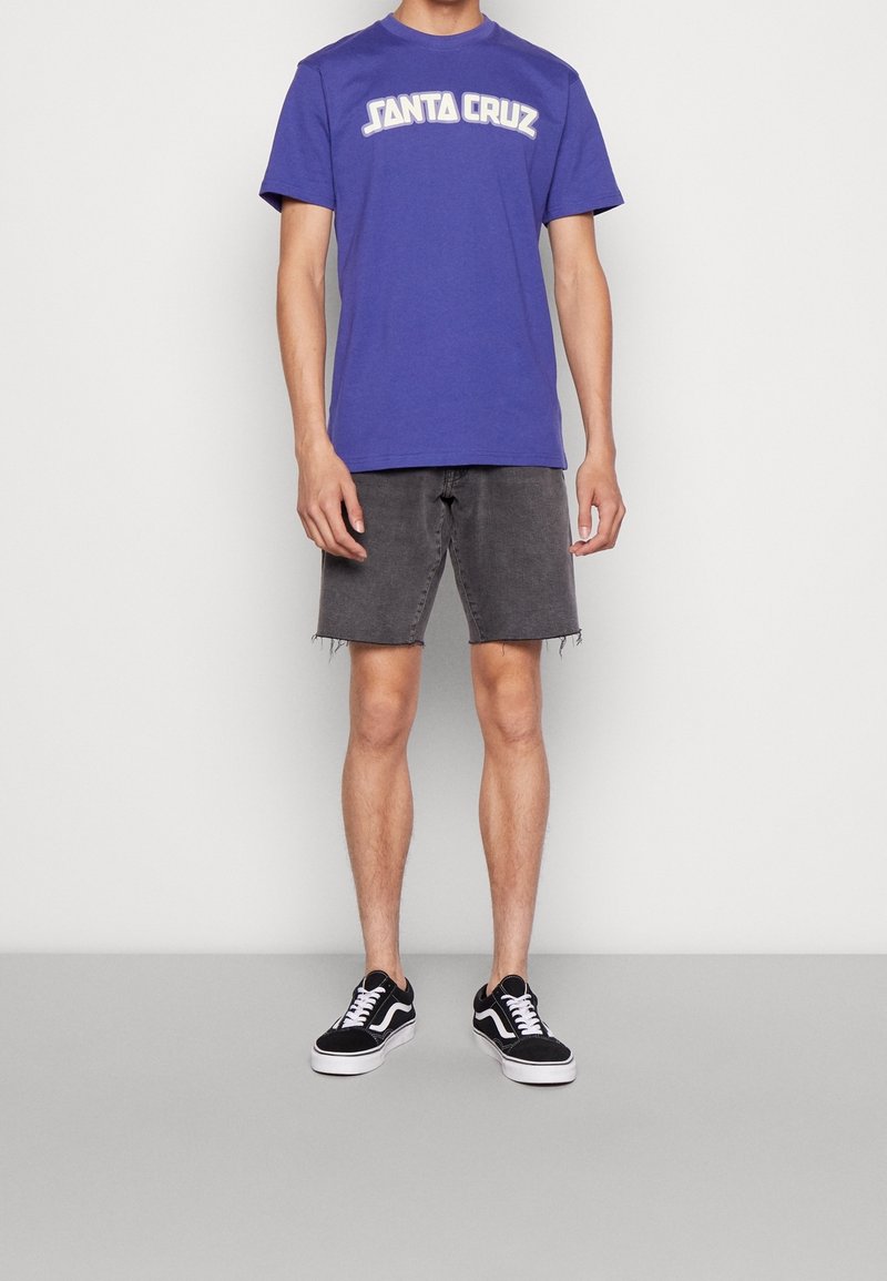 Lilla T-shirt med korte ærmer og "SANTA CRUZ" grafik, stylet med grå denimshorts og sorte Vans-sneakers med hvide detaljer.