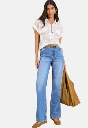 Woman wearing a white short-sleeve button-up blouse, high-waisted blue jeans, brown belt, brown flats, and holding a large brown tote bag.