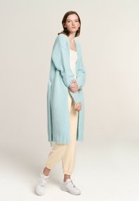 Light blue long cardigan made from soft knit material, featuring a relaxed fit, ribbed cuffs, and side pockets, worn over beige joggers and white sneakers.