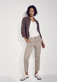 Pantalon beige, débardeur blanc et veste marron ample. Baskets blanches et grises, tissu lisse, coupe décontractée, pose en position debout contre un fond blanc.