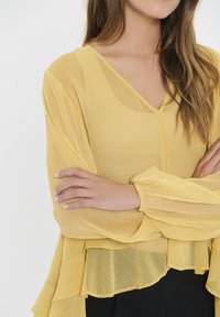 Geel gestructureerde blouse met een V-hals en een gerimpelde zoom, met lange, luchtige mouwen. De stof lijkt licht en doorschijnend te zijn.